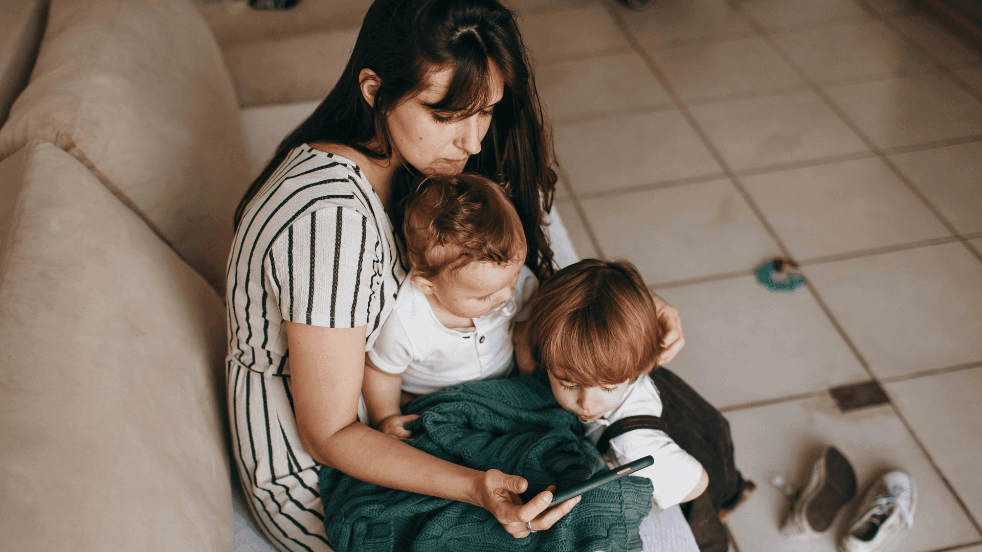 A mother sitting on a sofa with her two children on her lap, both looking at the phone she is holding, appearing intrigued by what they are watching.