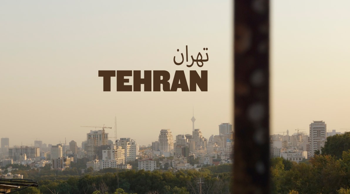Tehran skyline at hazy dusk with Milad Tower in the distance, framed past the out-of-focus carved stone column of a hilltop pavilion.