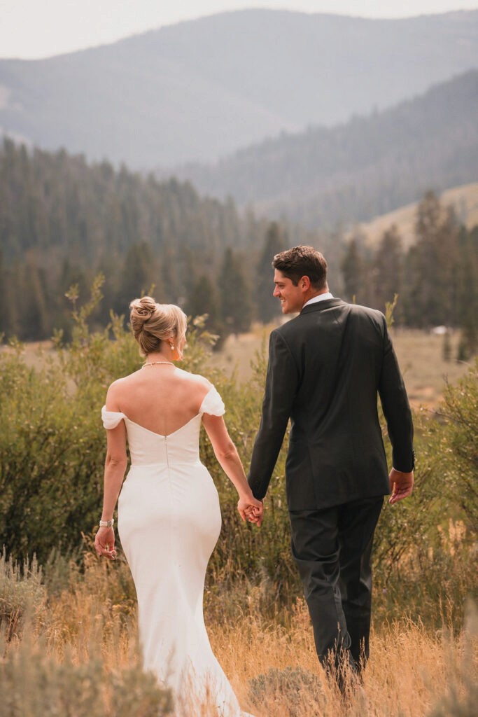 The bride and groom walk hand-in-hand away from the camera through tall grass toward a scenic vista of rolling hills and pine trees.