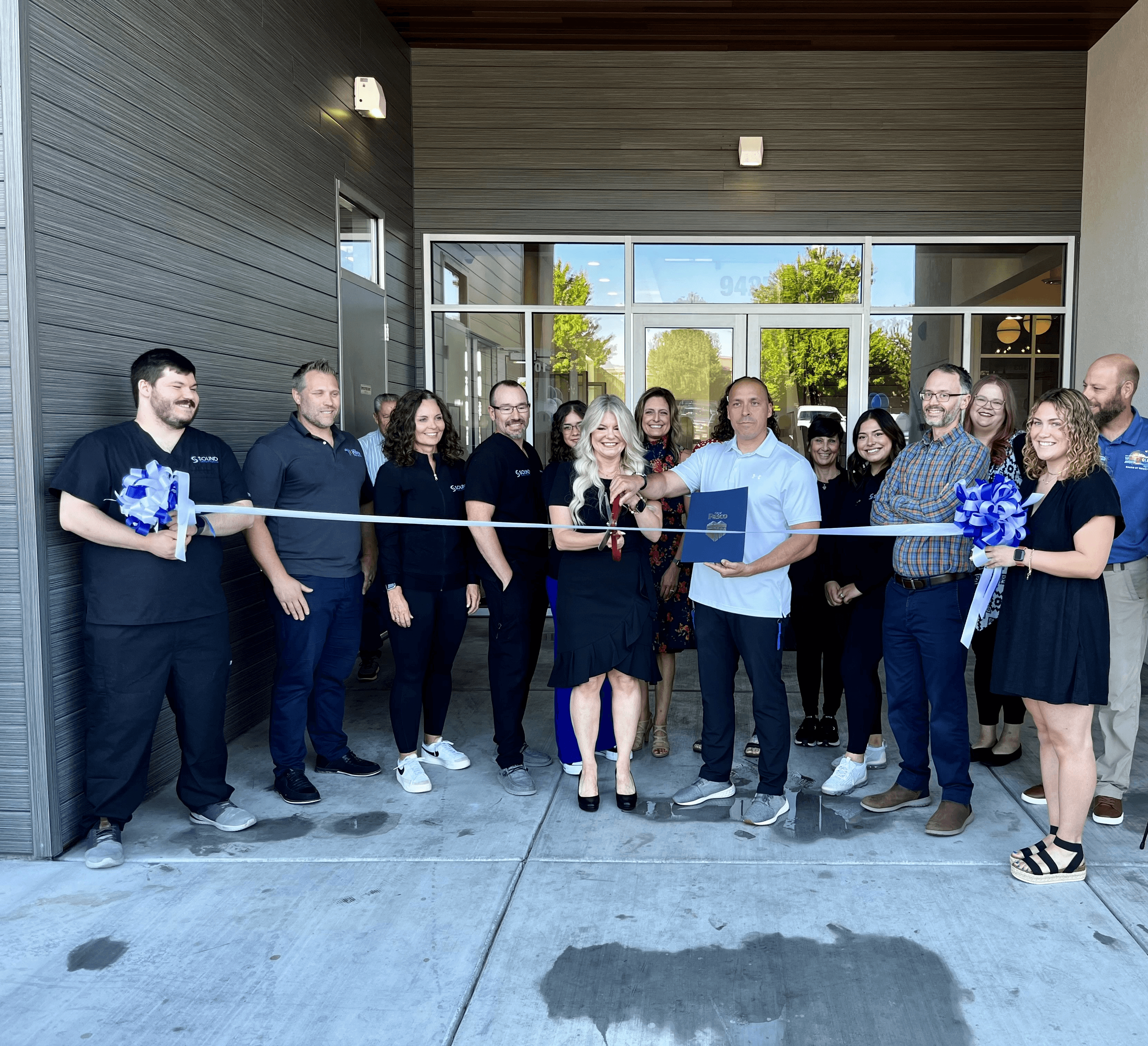 A group of people gathered outside a building, smiling and holding a ribbon for a grand opening ceremony.