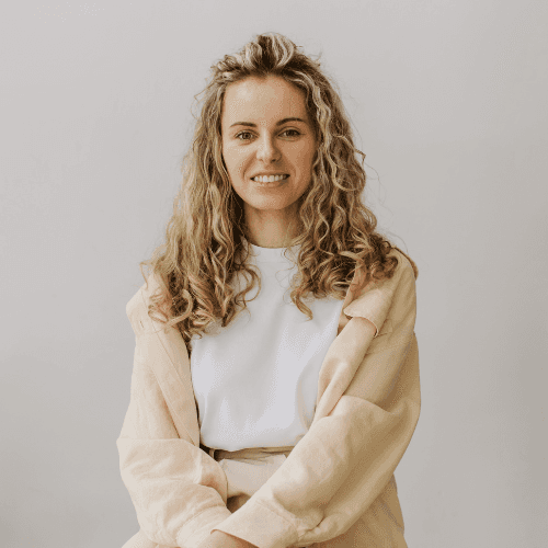 Smiling young woman with curly hair sitting at a laptop, in front of a teal wall.