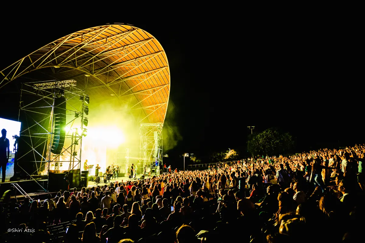 מבט פנורמי מרהיב על הופעה חיה של עידן עמדי תחת כיפת השמיים. הקהל הרב ממלא את האמפיתיאטרון מול במה מוארת בתאורה צהובה חמה ועוצמתית מתחת למבנה קשת ייחודי, מאת Shiriz Photography הצלמת שירי עציץ.