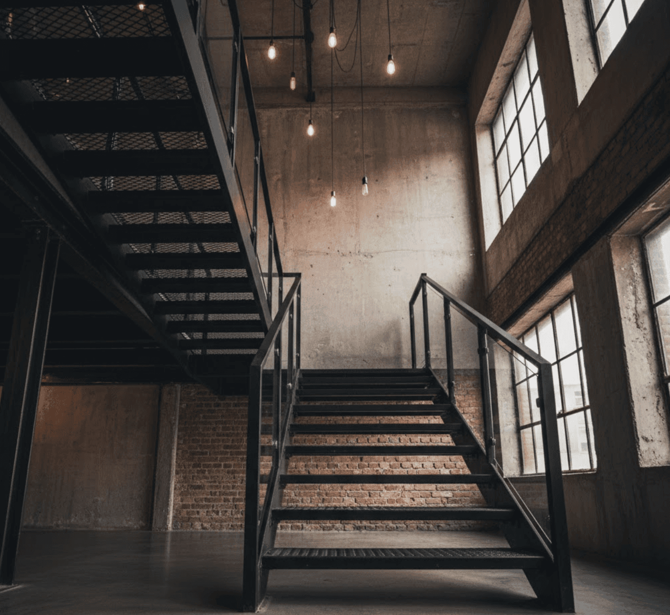 Industrial-style staircase with matte black powder-coated steel stringers and clear glass panels. Exposed steel I-beam structure, visible welds as design detail. Thick glass panels set in slim black steel frames. Raw concrete wall backdrop with exposed brick accent. Black steel mesh landing platform. Edison bulb pendant lighting. Converted warehouse loft space with high ceilings and large factory windows. 