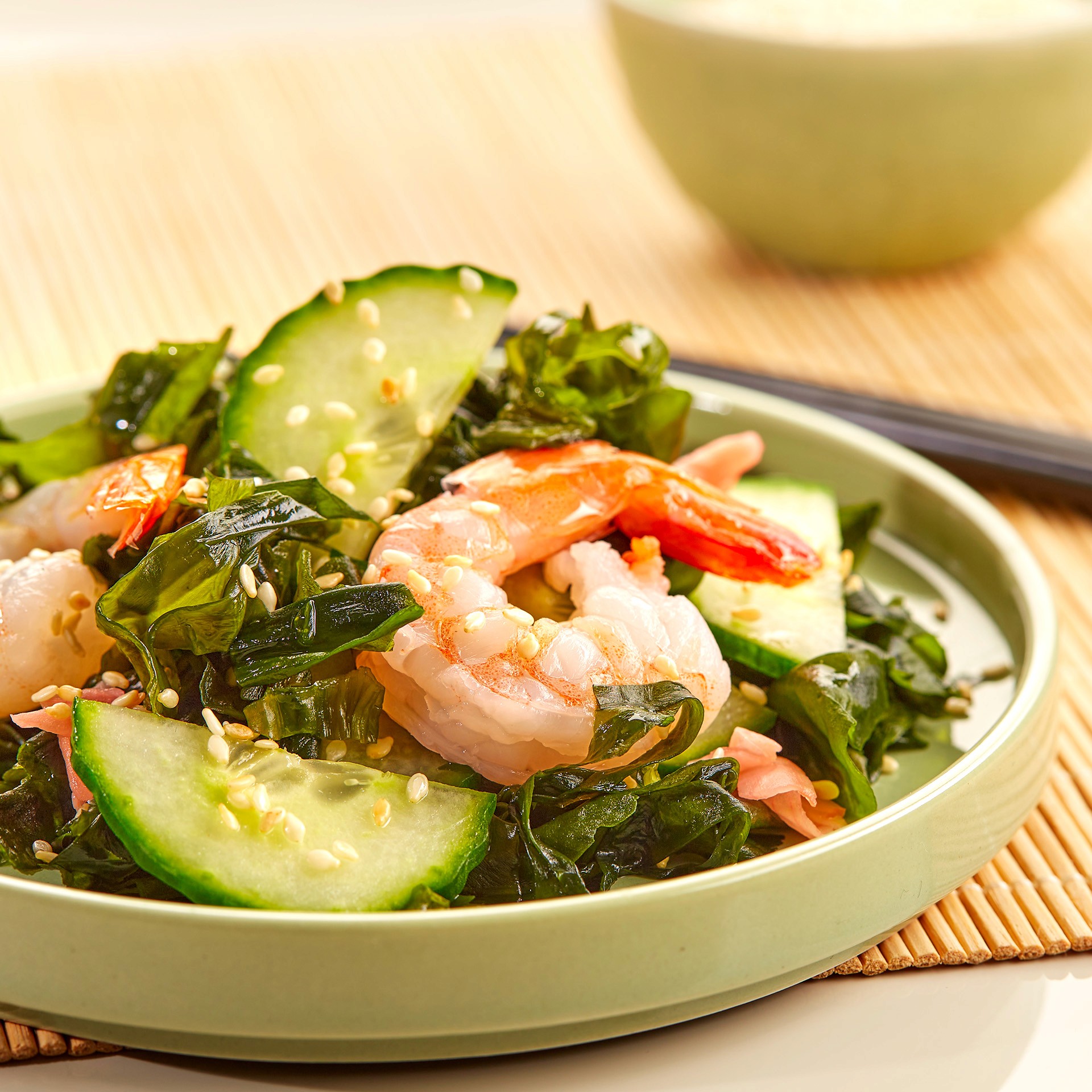 A light green plate features a fresh seaweed salad topped with plump shrimp, sliced cucumbers, and garnished with sesame seeds, set against a bamboo mat background with a blurred pale green bowl in the distance.