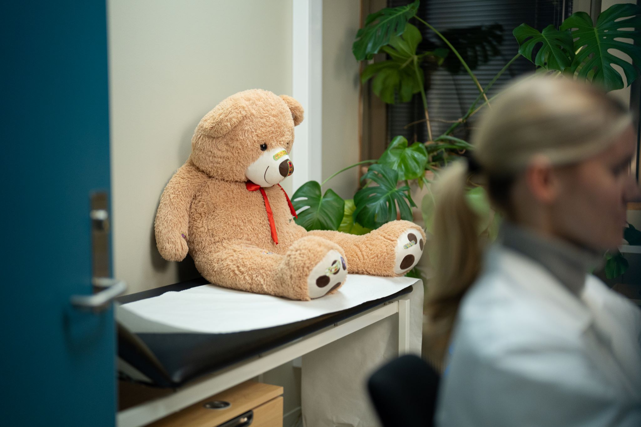 En bamse sitter på et skrivebord ved siden av en person som jobber ved en datamaskin på et fastlegekontor.