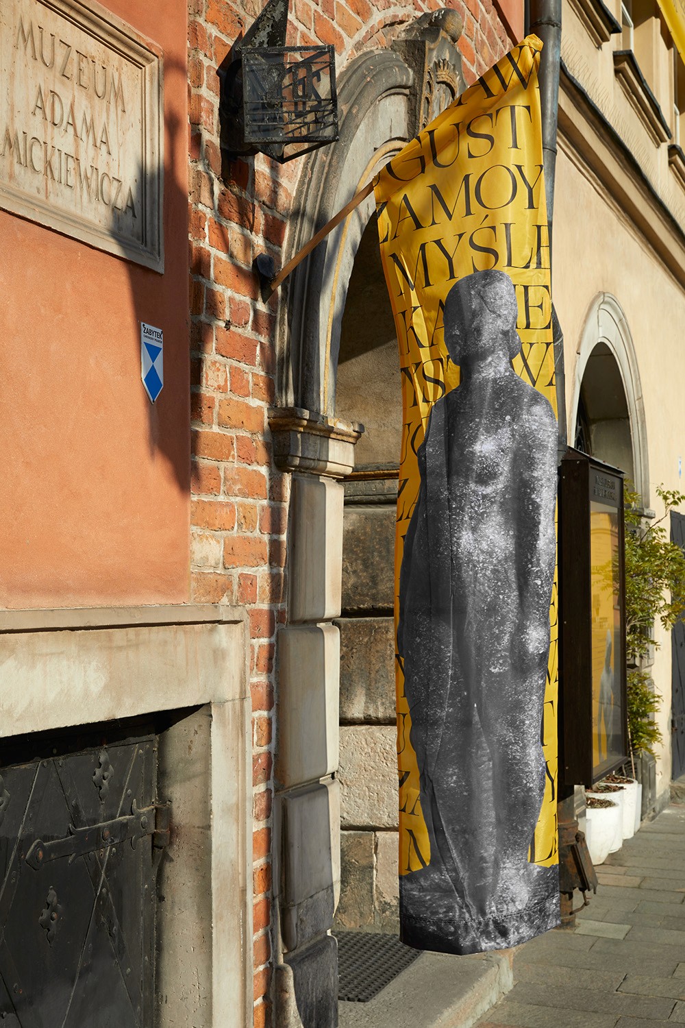 A large, bright yellow fabric banner featuring bold typography and a grayscale sculptural image hanging from a historic brick building. This showcases the studio's proficiency in environmental graphic design and large-scale institutional signage.
