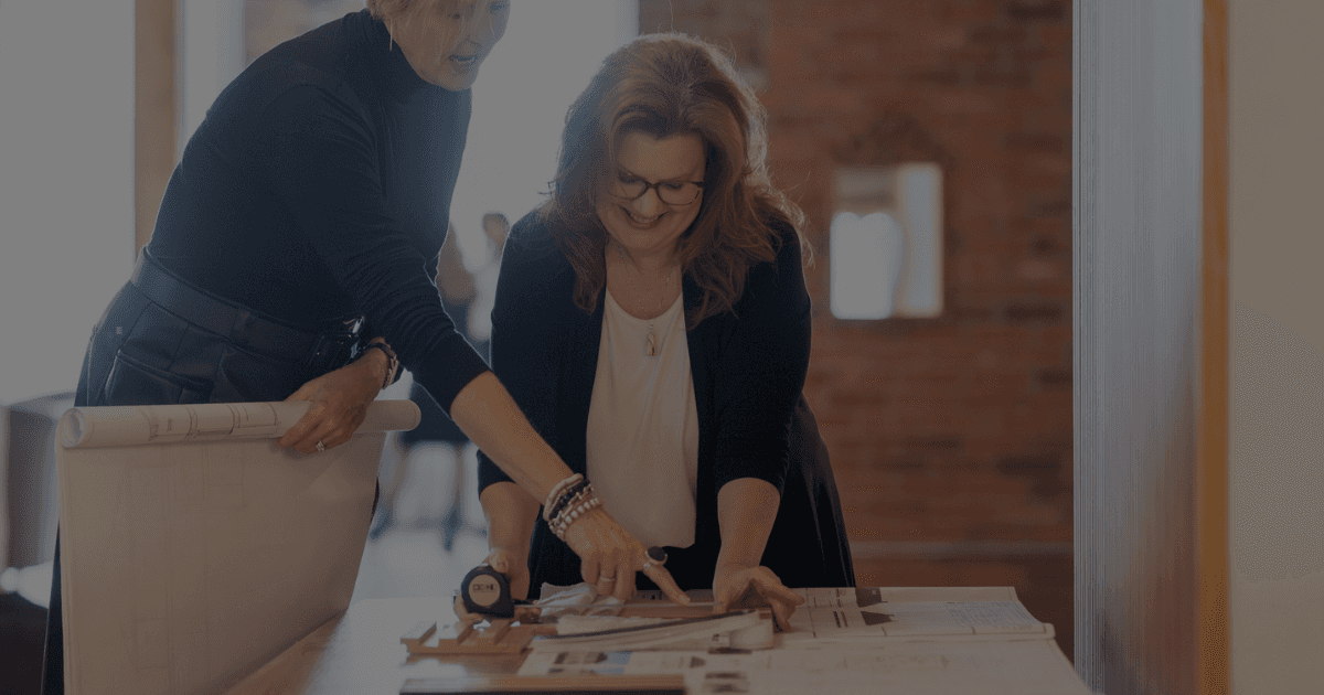 Two designers look at a set of drawings and samples. One designer points to a specific area on the drawing. Both are females with blond hair and fair skin wearing dark clothing.