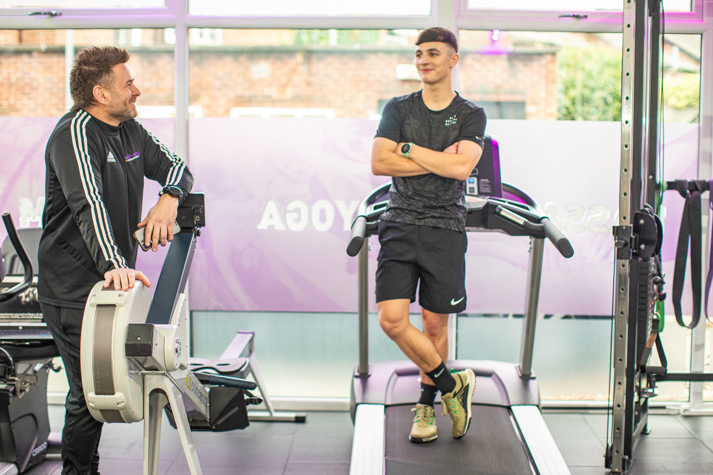 Rufus doing a fitness consultation to identify a realistic programme for strength training and muscle building. coach is smiling with his arms crossed, wearing a black 