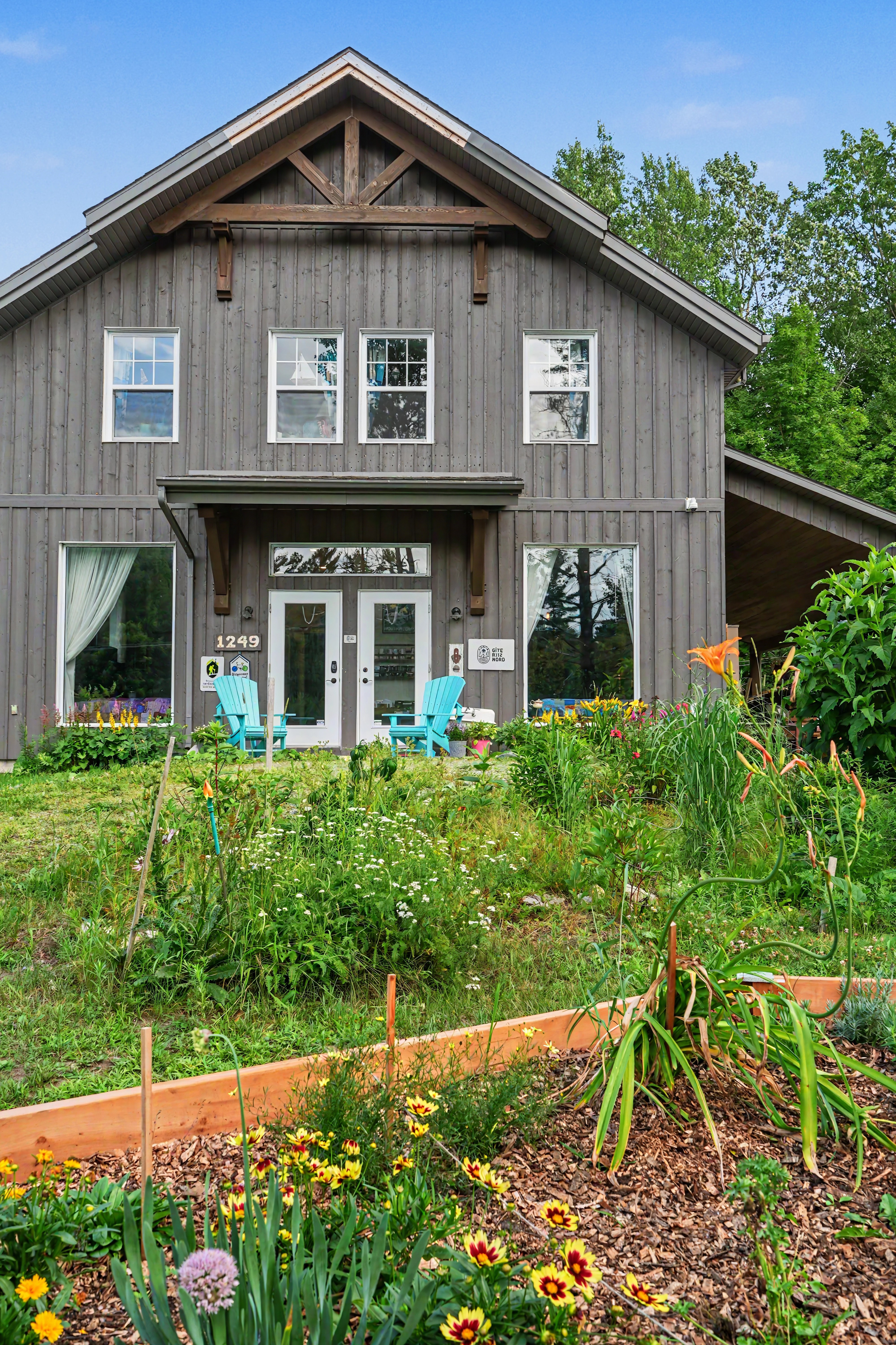 Façade en bois foncé du gîte R112 Nord entourée de fleurs sauvages colorées, par une journée d’été ensoleillée.