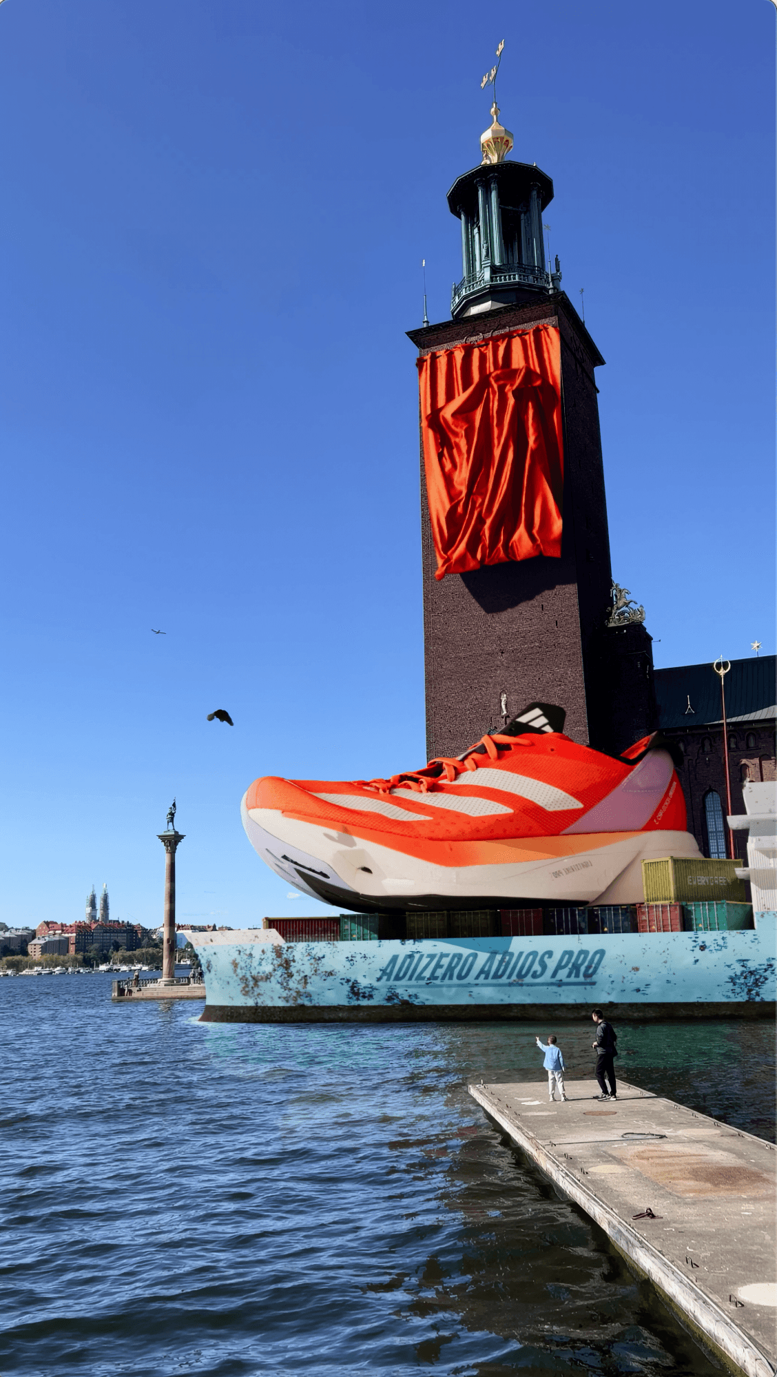 A massive orange Adidas sneaker transported by a cargo ship past a city tower.