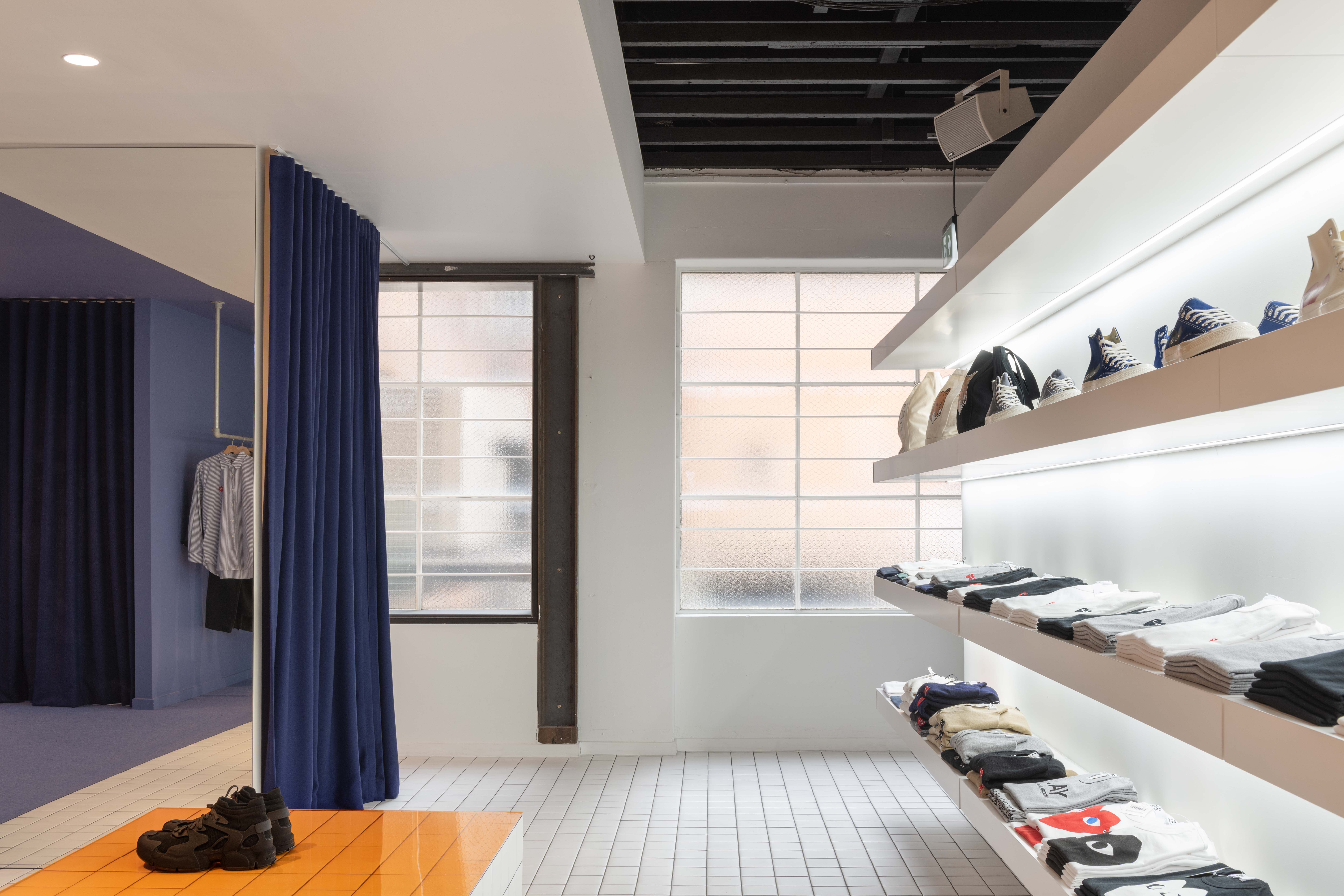 Bright retail area inside the 199A® space, featuring floating wall shelves with folded garments and shoes, tiled flooring, large industrial windows, and a blue-curtained fitting zone.