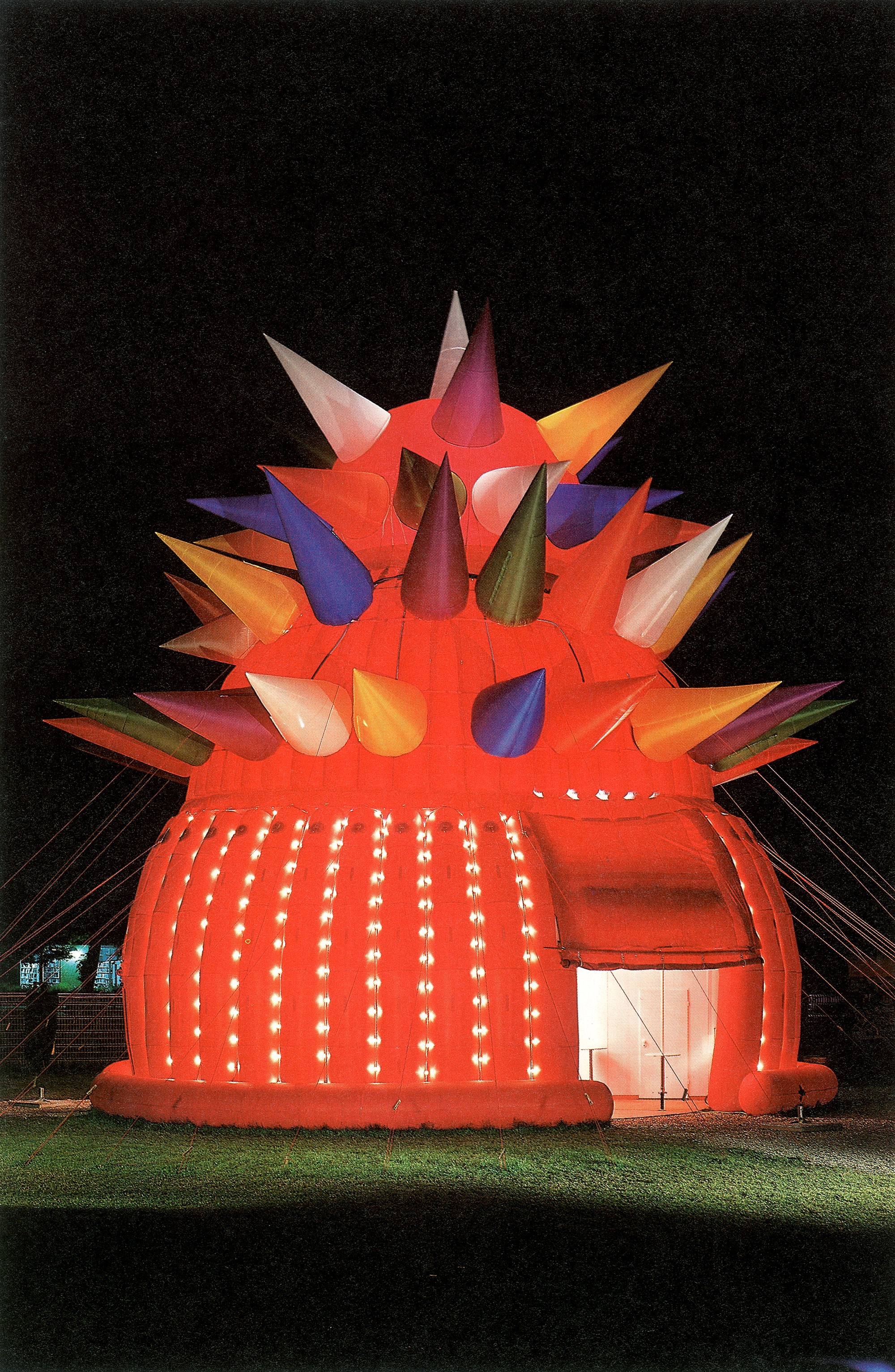 Aufblasbare rote Kuppel mit unterschiedlich farbigen, spitzen Elementen auf der Oberfläche und beleuchteter Fassade bei Nacht.