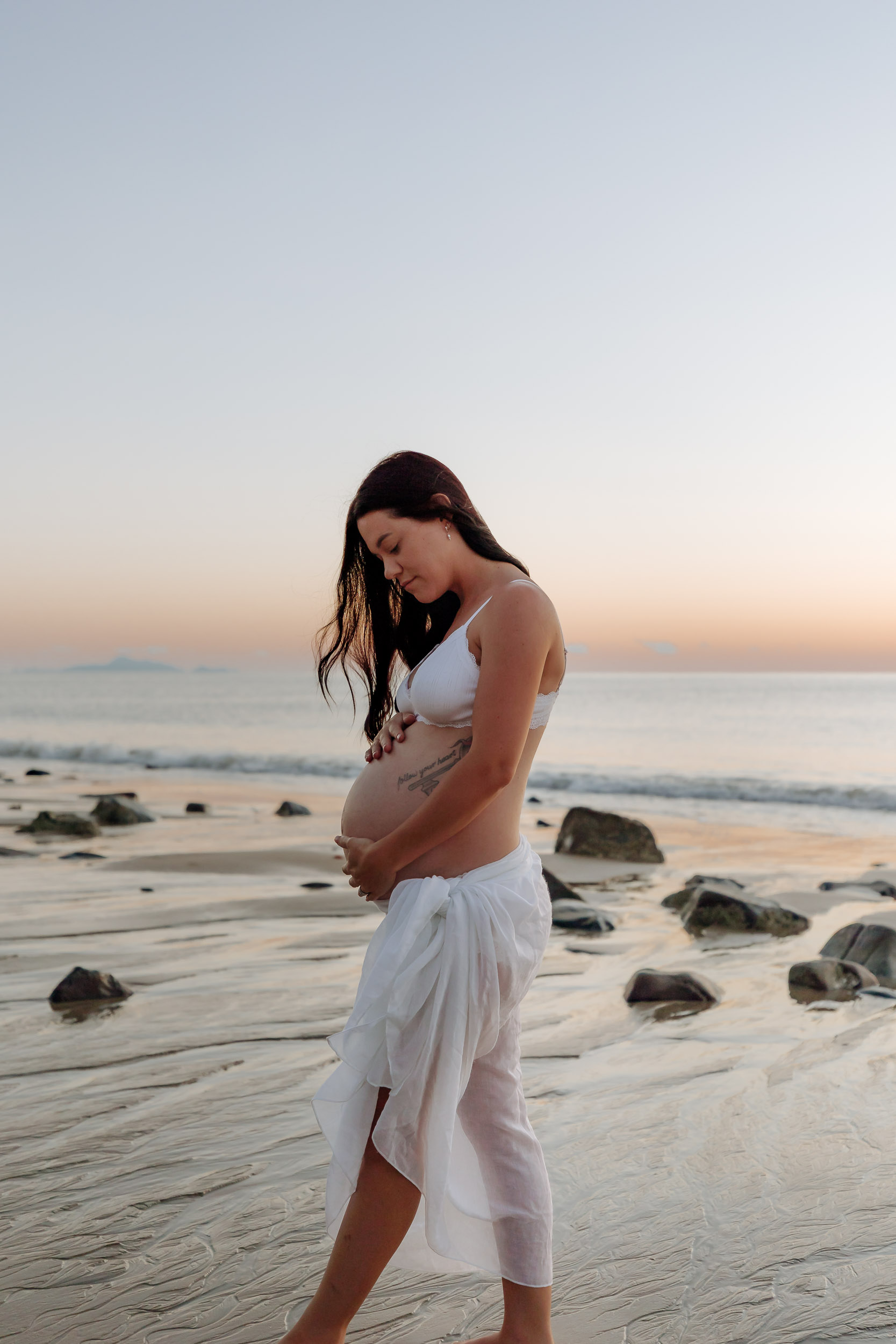 Expectant mother photographed alone at sunrise, walking along the shoreline with gentle waves and pastel skies creating a calm, ethereal coastal backdrop.