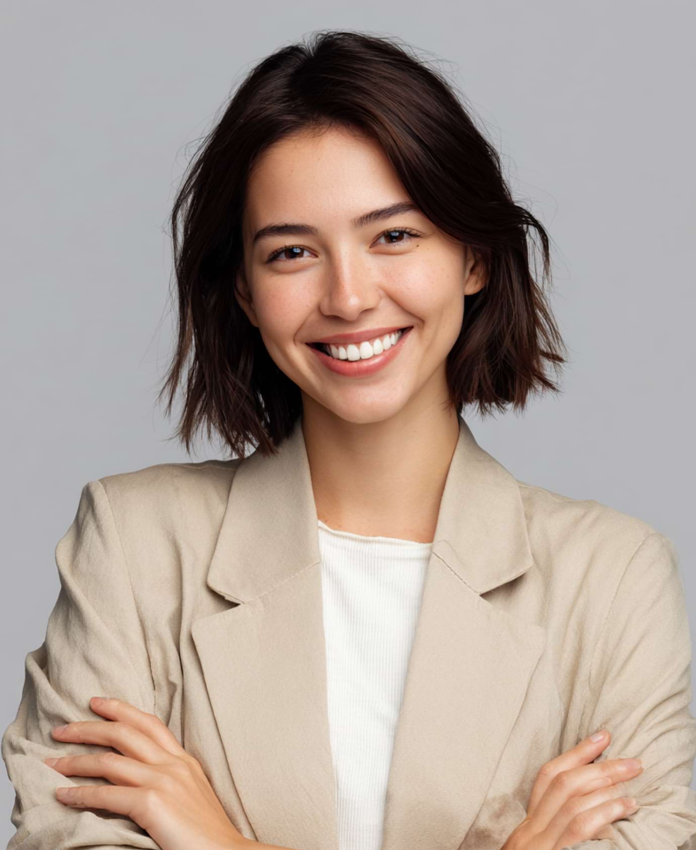 Smiling woman with short brown hair, wearing a beige jacket and white shirt, stands with arms crossed against a grey background, conveying confidence.