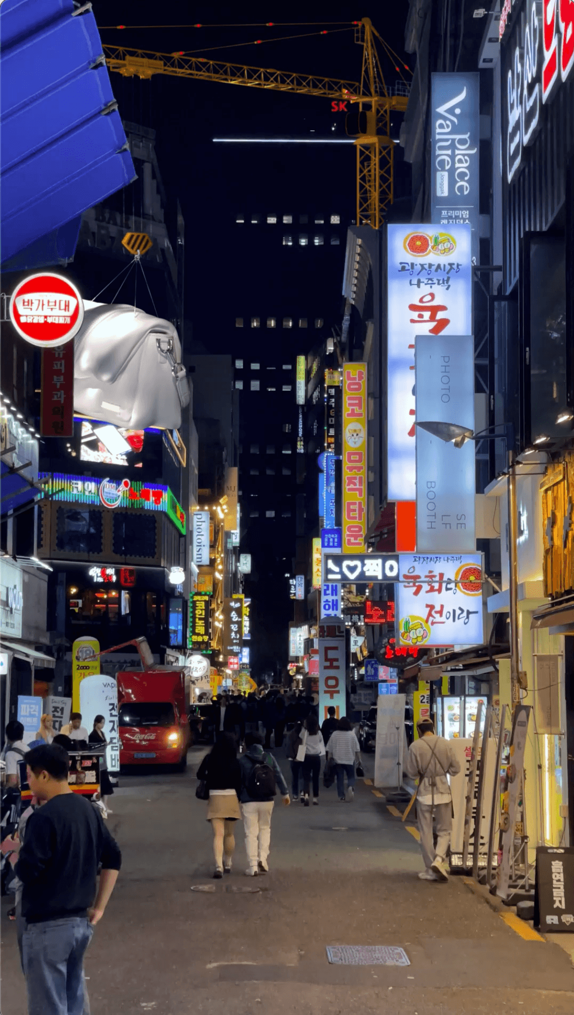 A massive white Jacquemus bag suspended by a crane over a bustling neon-lit street in South Korea