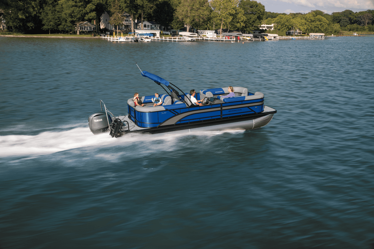 A blue pontoon boat with a powerful outboard motor cruises smoothly across a scenic lake, surrounded by lush green trees and quaint waterfront homes under a clear blue sky.