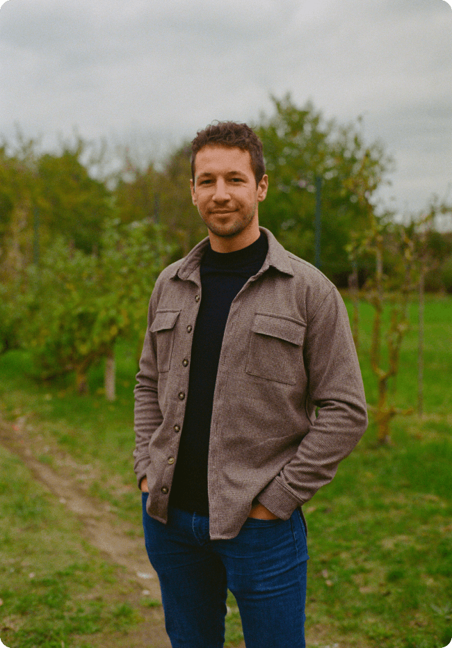 Portrait d'un homme souriant en chemise marron et jean, posant avec les mains dans les poches dans un jardin verdoyant.