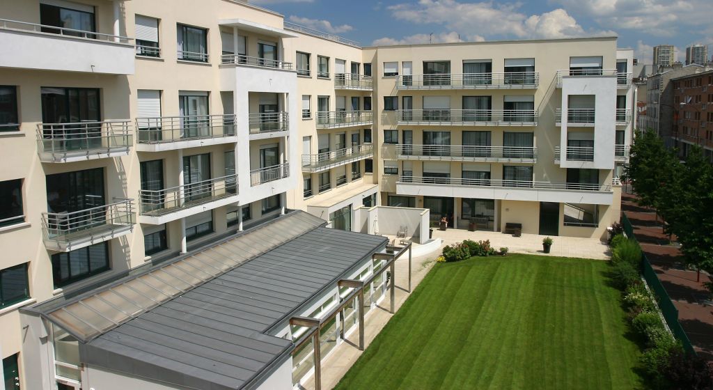 Modern multi-story clinical facility exterior with white and beige cladding, green manicured lawn, and paved courtyard