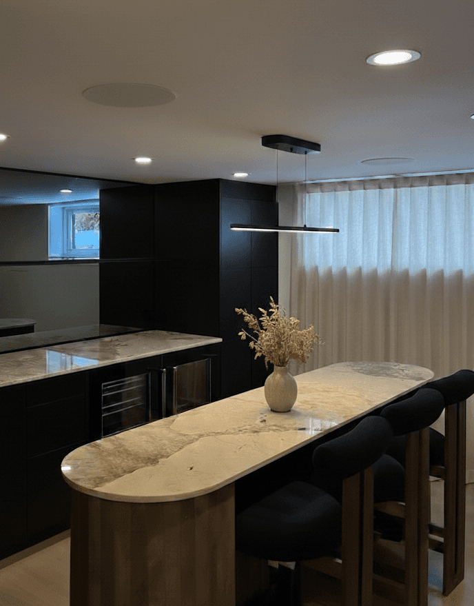Voici un élégant coin bar au design contemporain dans lequel on retrouve un îlot aux courbes douces en Dekton, s'harmonisant parfaitement avec des armoires noires mates, des tabourets foncés, un luminaire minimaliste et de légers voilages qui diffusent une lumière douce et chaleureuse.