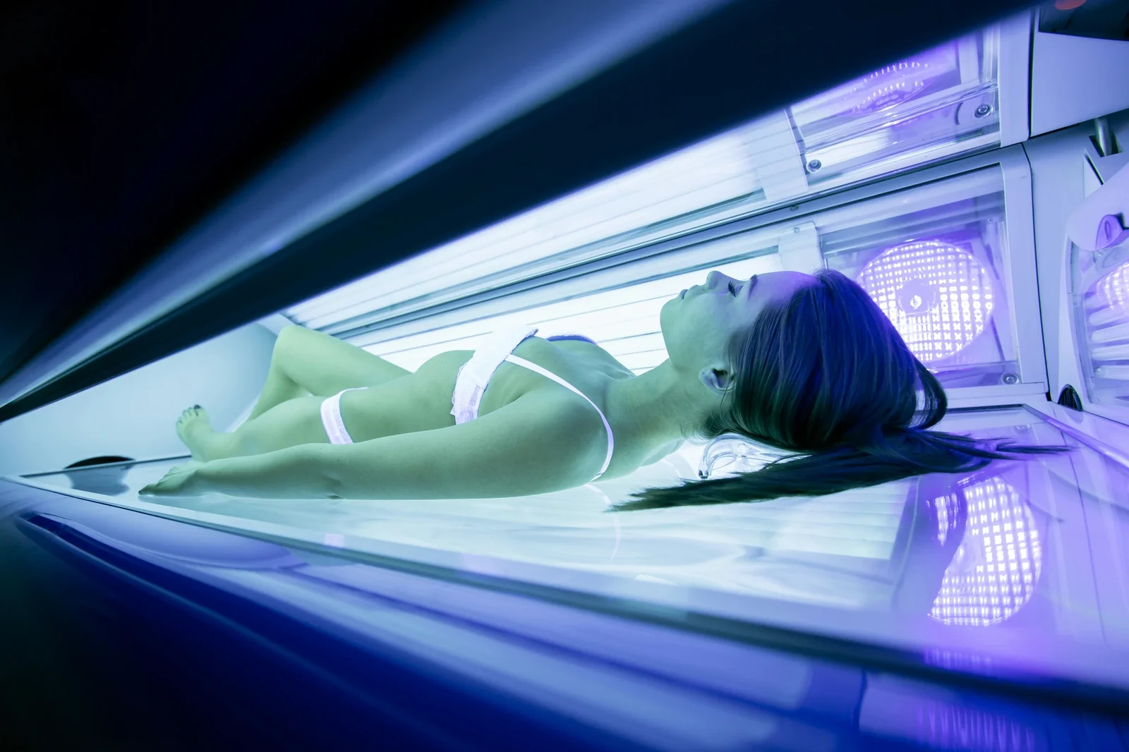 Woman lying in an open tanning bed with purple UV lights illuminated inside the salon equipment.