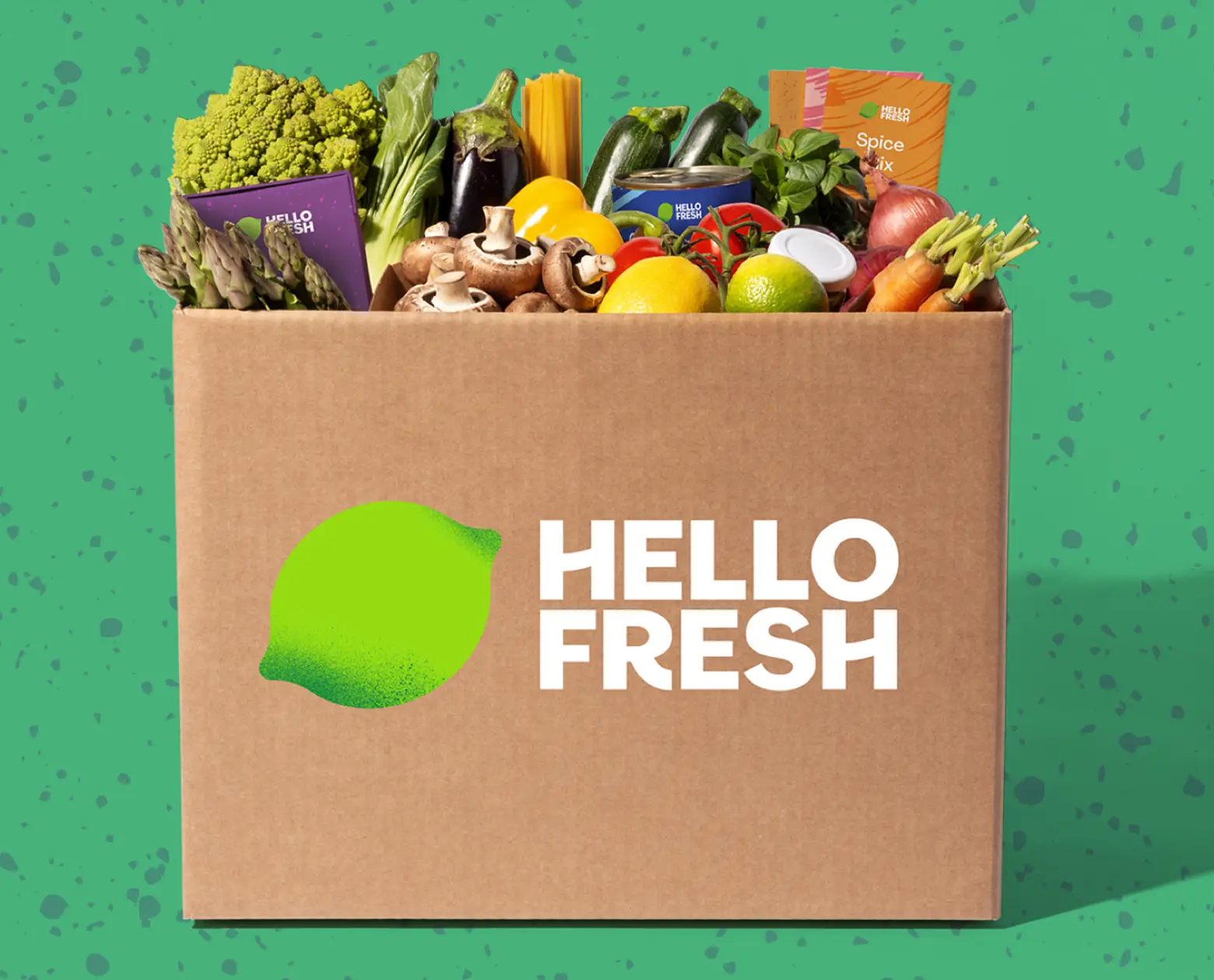 A close-up view of an open HelloFresh cardboard box brimming with vibrant, diverse fresh produce including romanesco broccoli, asparagus, and bell peppers against a green background.