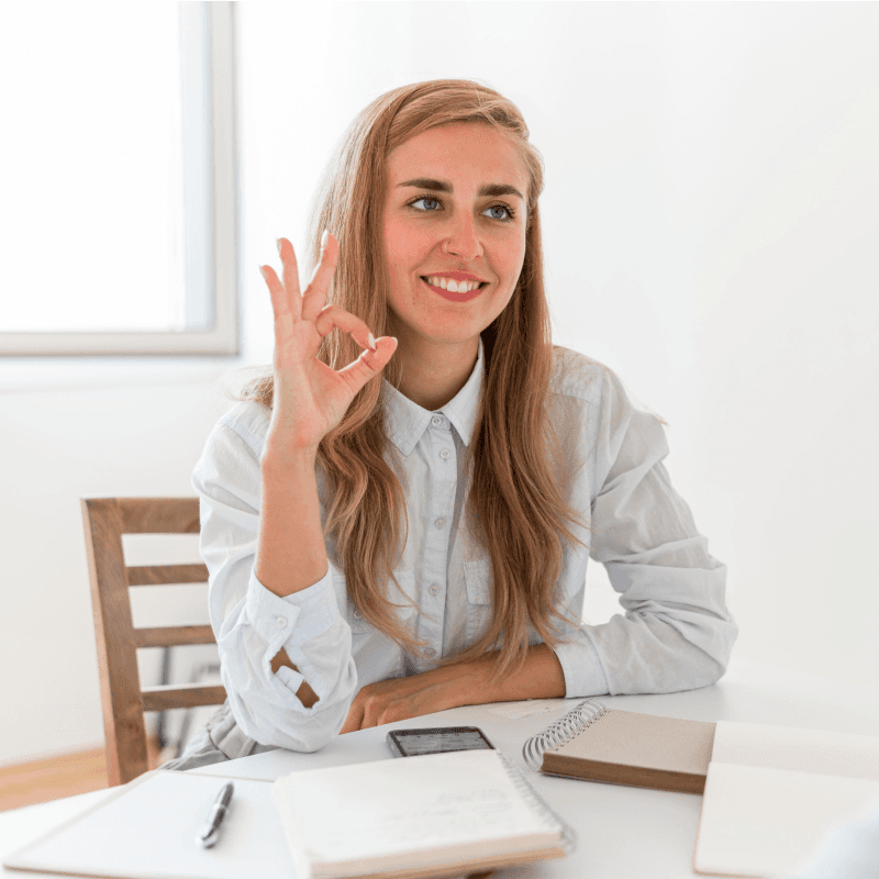 A smiling woman with long blonde hair sits at a desk with notebooks and a smartphone, making an OK hand gesture. She wears a light-colored shirt and is in a bright, minimal room.