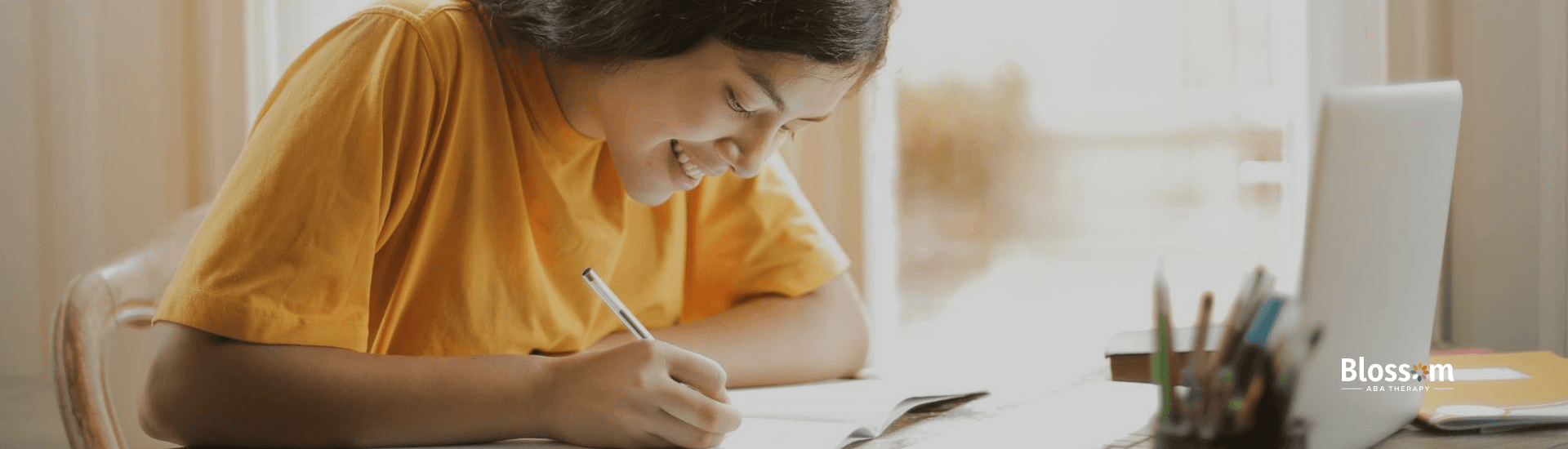 A BCBA aspirant writing in a notebook while smiling, seated at a desk with a laptop and stationery.