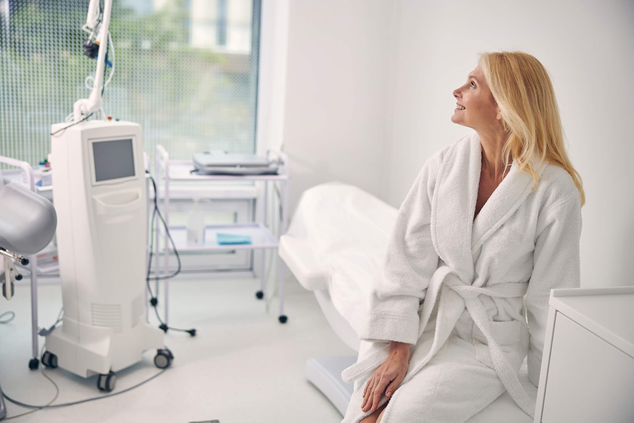 Woman in a white robe sitting in a medical clinic, looking toward equipment and a healthcare professional out of frame