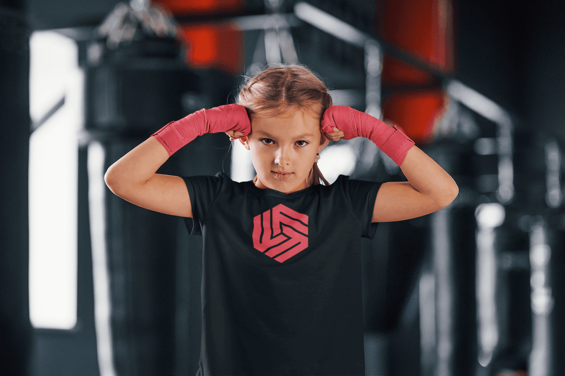 A young girl standing confidently in a boxing gym, wearing a black T-shirt with the 'Flexibility For Fighters' logo in red on the front. She has her hair in braids and is wrapping her hands with pink hand wraps, positioned as if ready to start a training session. The background is blurred, showing the gym’s punching bags and equipment, adding to the energetic and focused atmosphere.