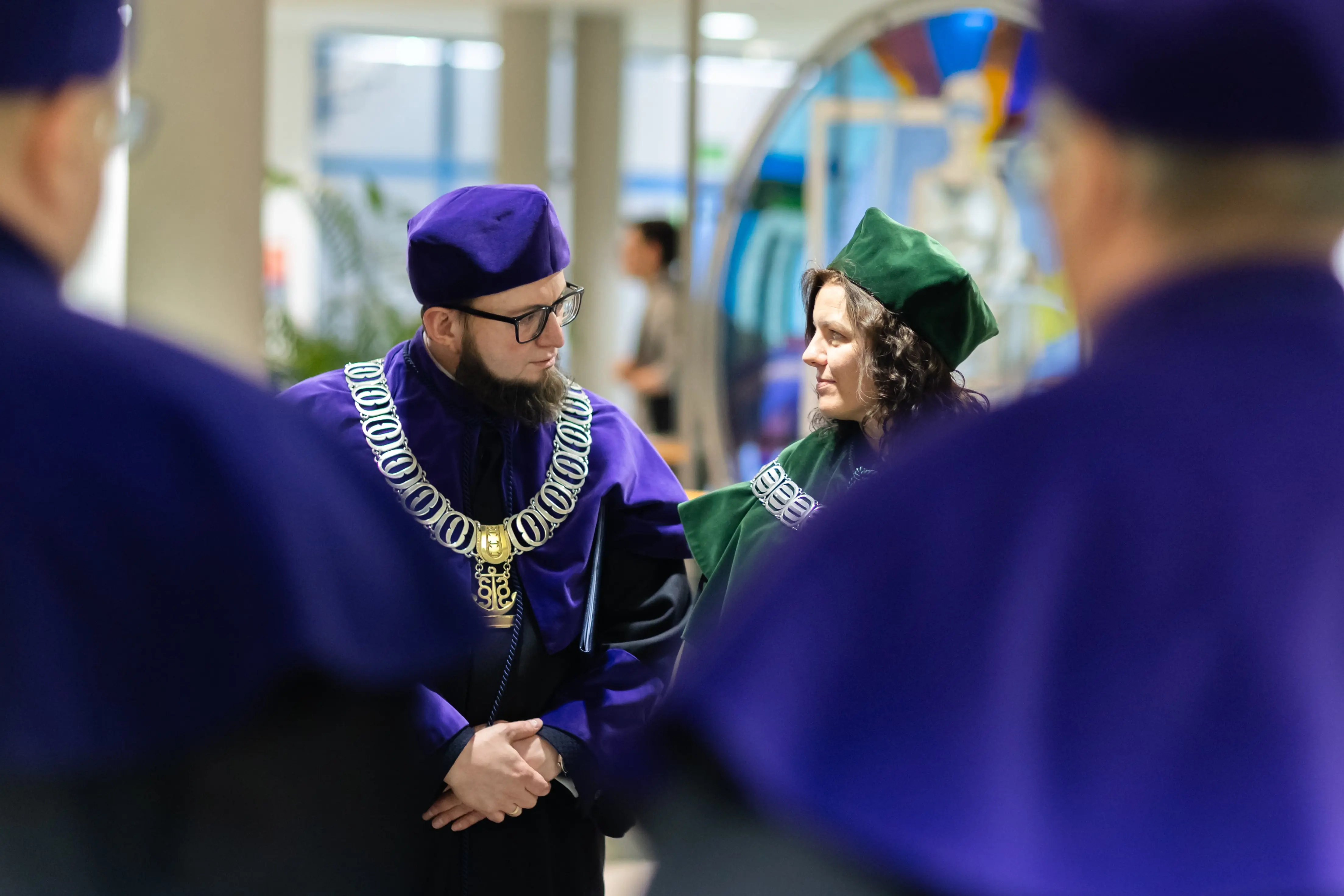 Dwoje pracowników akademickich ubranych w tradycyjne stroje, z ozdobnymi łańcuchami na szyi, rozmawia w tle, podczas uroczystości akademickiej. Widać również sylwetki innych osób w podobnych strojach.