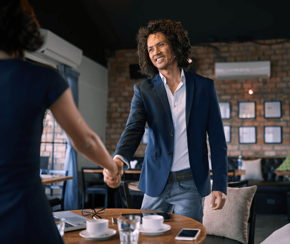 A man in a blazer shakes hands with a woman in a restaurant setting, both smiling and engaged in conversation.