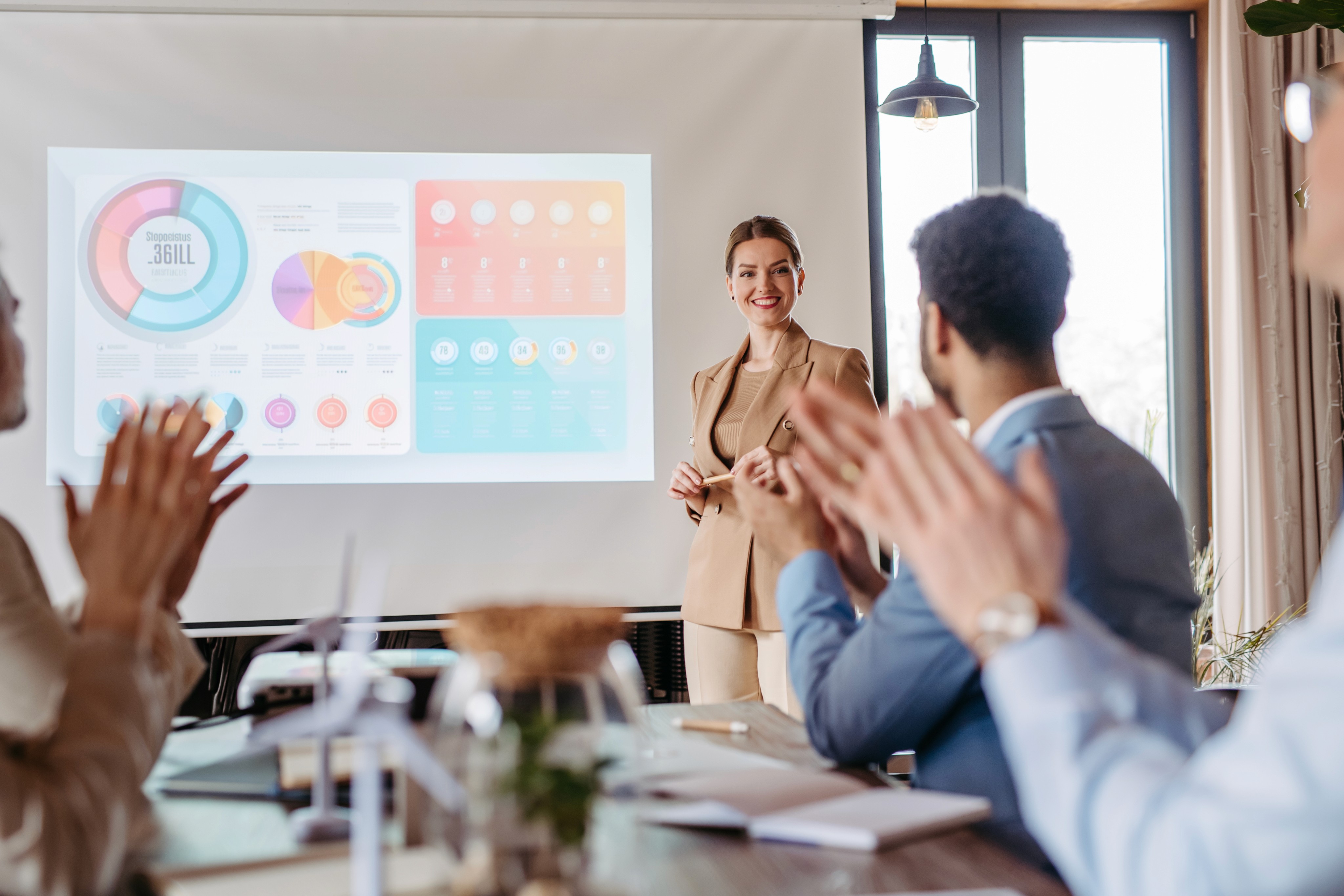 Woman in tan suit presenting data to a room of colleagues in a board meeting