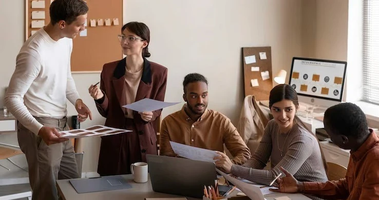 Equipe em volta de uma mesa com laptop e papéis, com uma mulher de pé apresentando