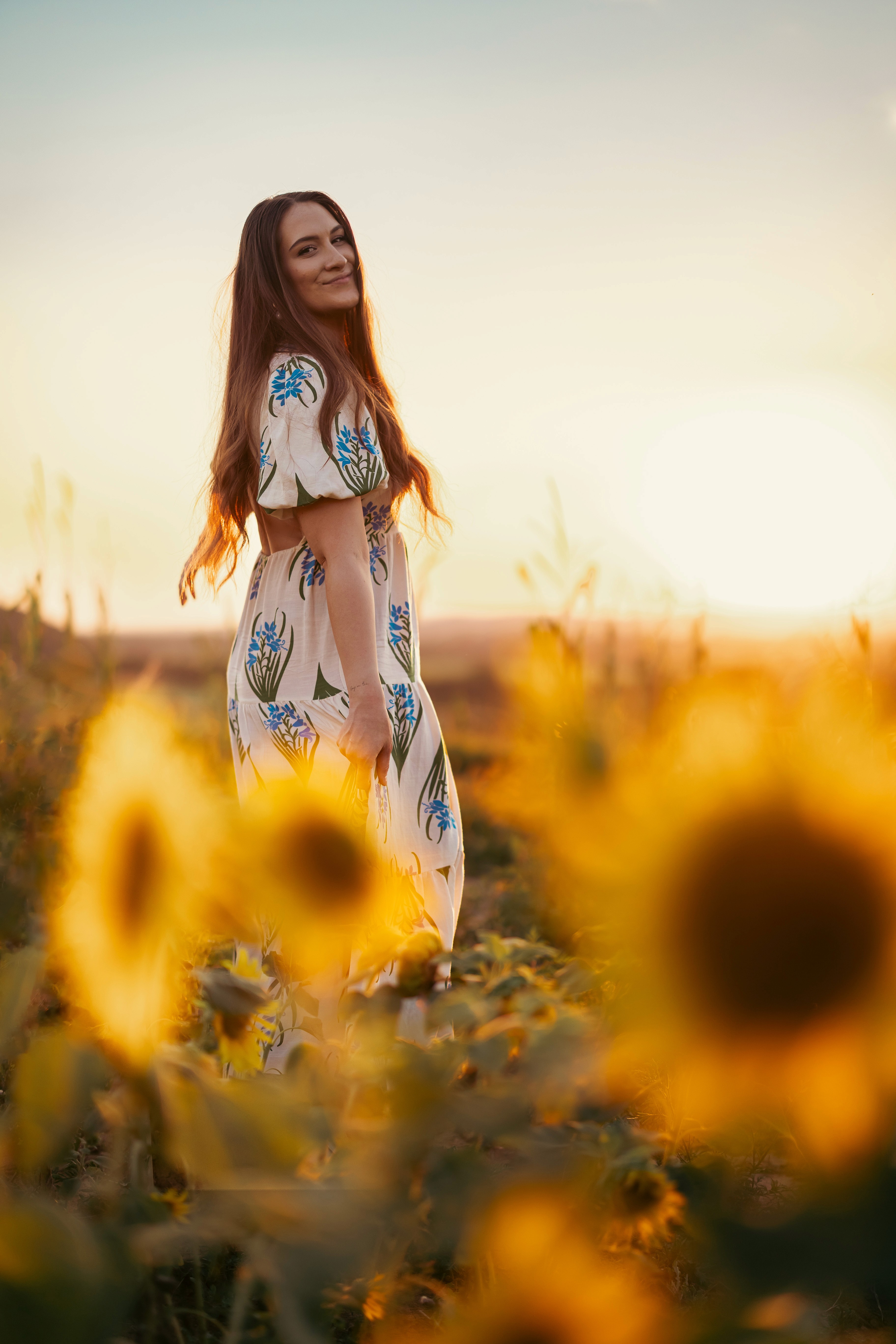 Fiatal nő áll napraforgómezőben naplementekor, virágos ruhában mosolyogva, hosszú barna haja szabadon hullik a vállára.