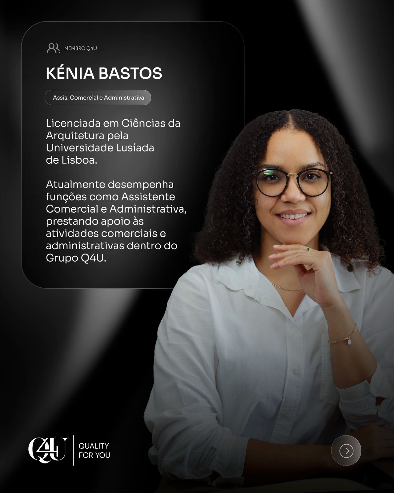 A woman with curly hair and glasses smiles warmly, wearing a peach dress. She's in a sunlit room filled with lush green plants, creating a cozy, serene atmosphere.