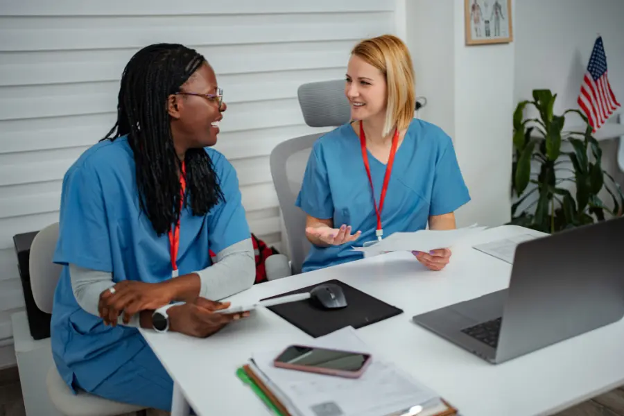 AI nurse communication between two nurses in blue scrubs laughing and reviewing documents together at an office desk with a laptop and American flag in the background.