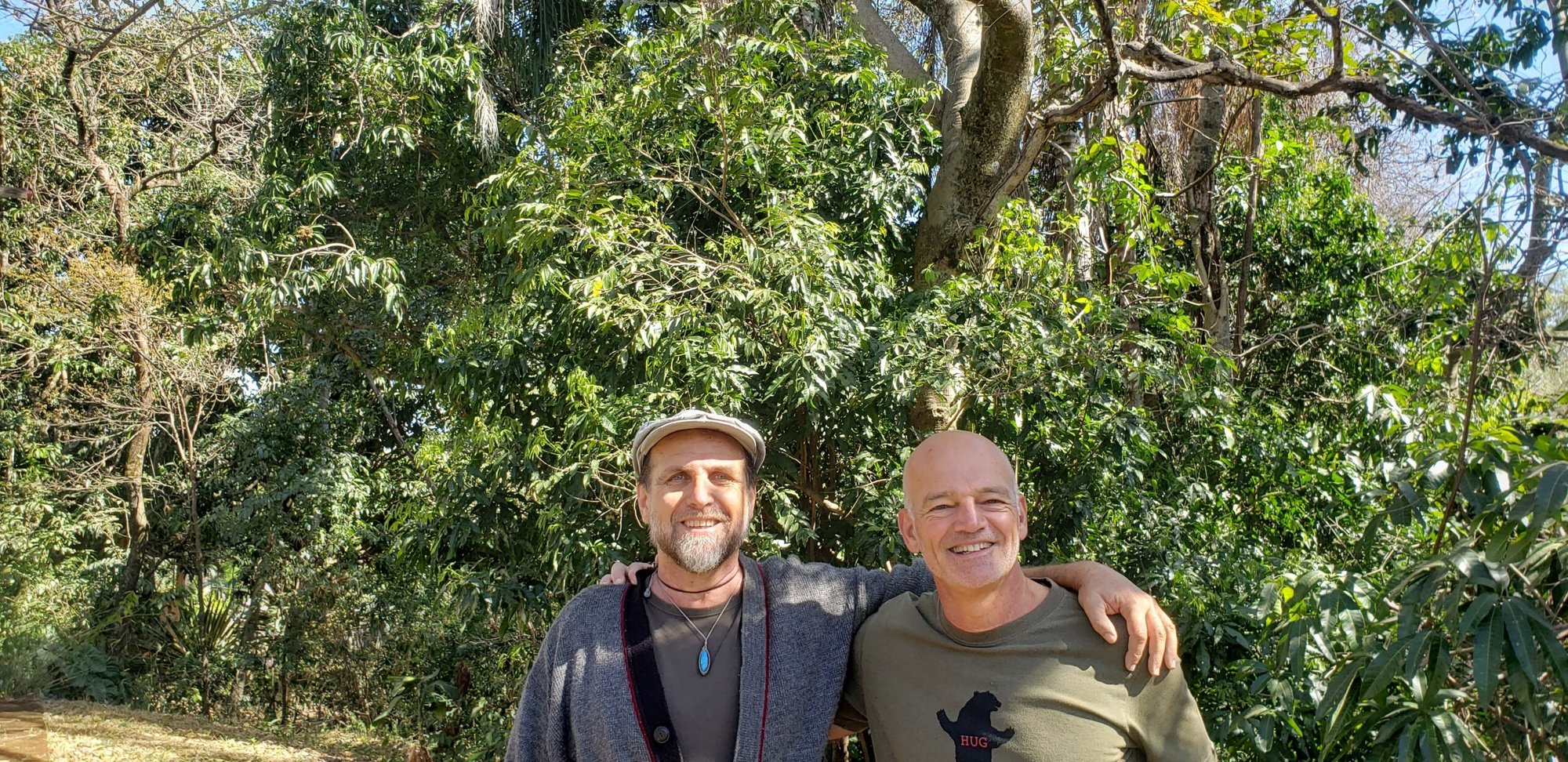 Dois homens abraçados e sorrindo em frente à vegetação da mata em dia ensolarado