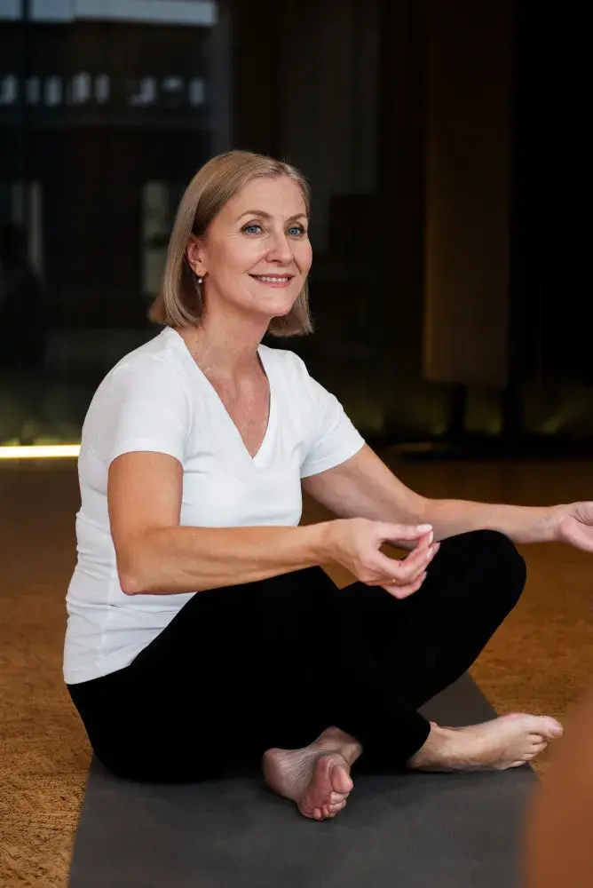 A woman with short, blonde hair sits cross-legged on a yoga mat in a calm, indoor setting, wearing a white t-shirt and black pants, appearing relaxed and content.
