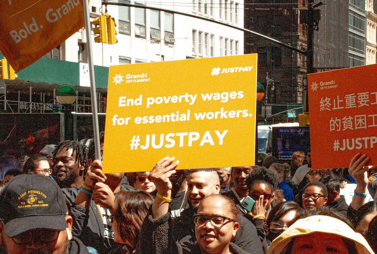 A crowd of people with the image focused on a sign that says "end poverty wages for essential workers"