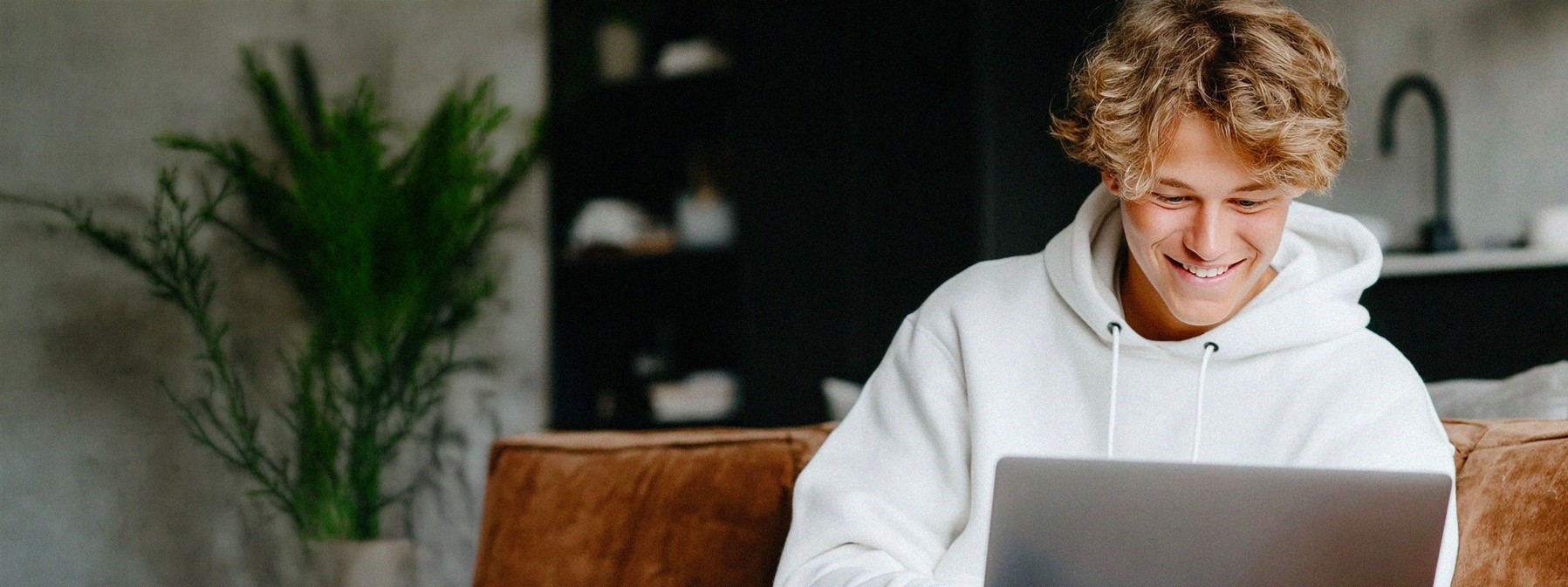 A young person with short, curly hair sits at a desk working on a computer in a bright classroom or office setting. They appear focused on the screen, with other people and desks softly blurred in the background, suggesting a collaborative learning or work environment.
