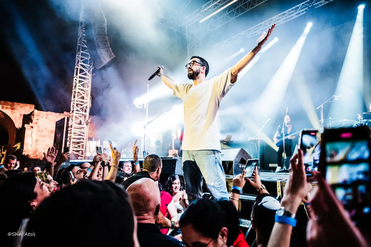 צילום מרגש מתוך הופעה חיה של חנן בן ארי, האמן עומד על קצה הבמה בידיים פרושות מול קהל מעריצים שמצלם ושר איתו יחד באווירה מחשמלת, מאת Shiriz Photography הצלמת שירי עציץ.
