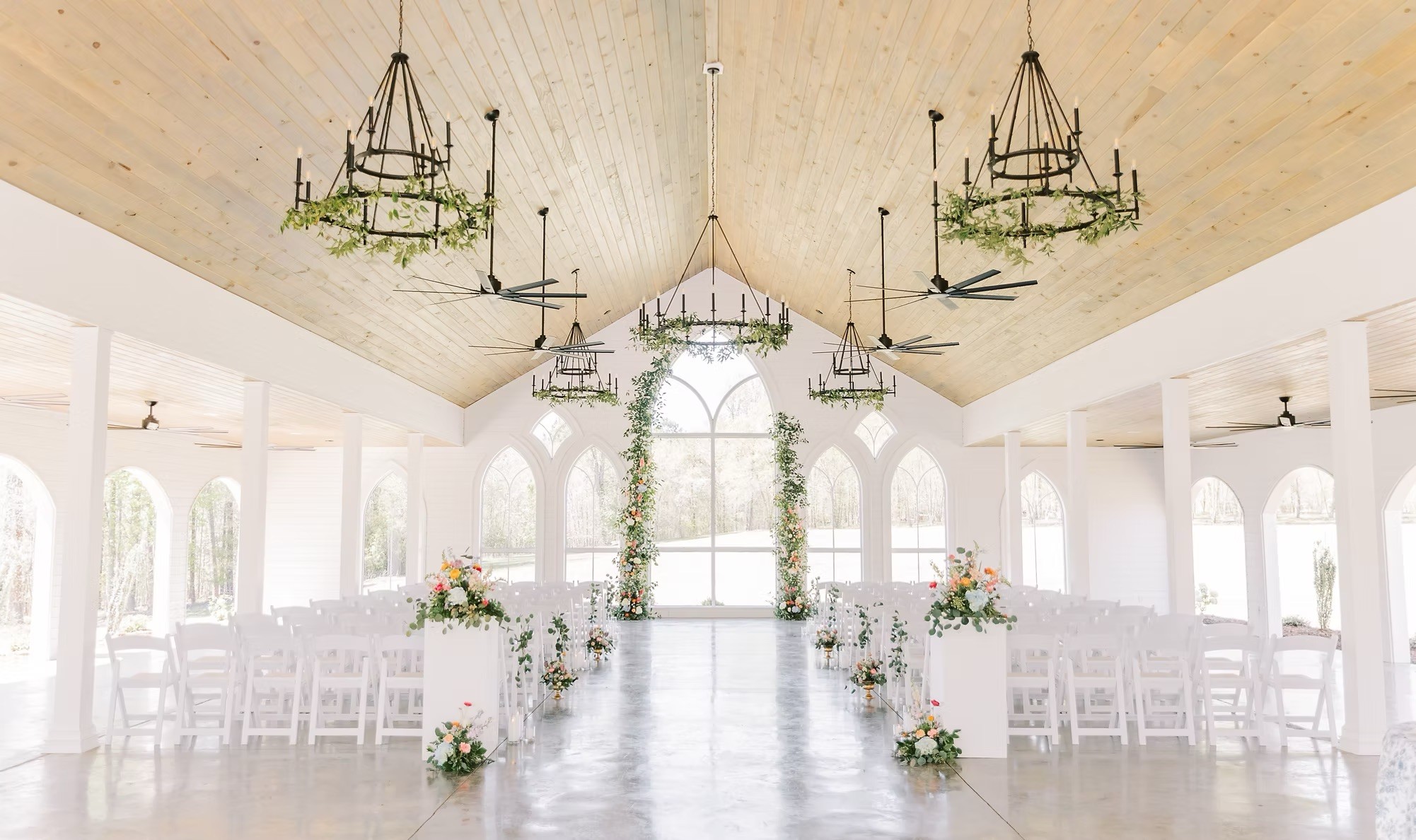 A serene outdoor wedding setup with white chairs arranged on green grass under a canopy of trees.