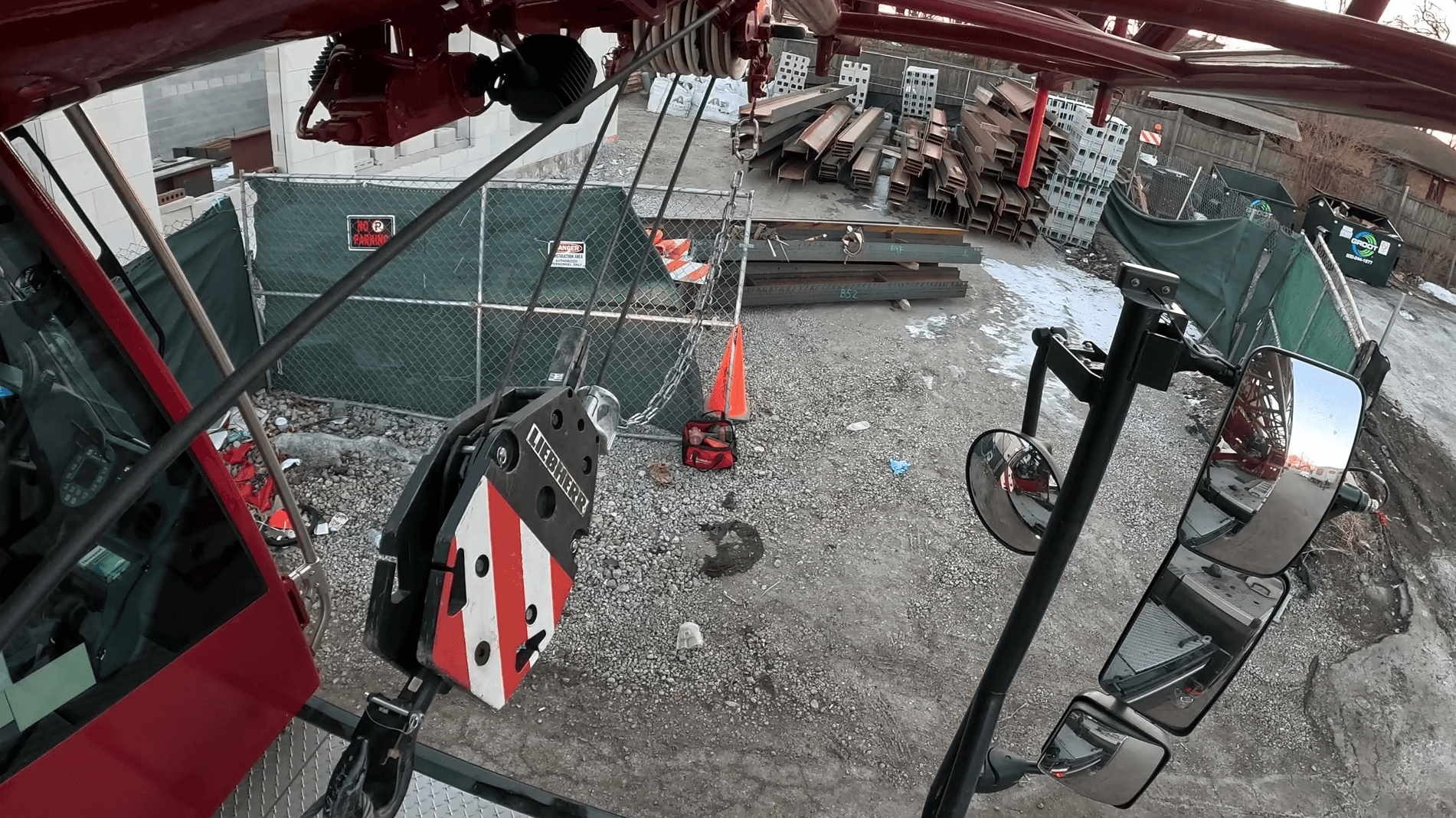 A construction site view from the inside of a crane cab, showing the crane's hook and cables lifting materials, with a backdrop of building supplies, safety cones, and a fenced-off work area.