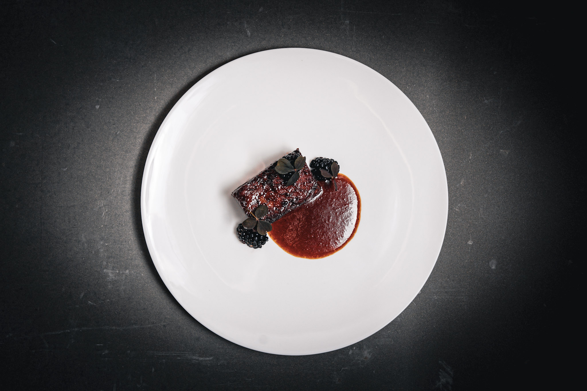 Beef short rib with reduced sauce and whole blackberries, plated simply with minimal garnish and dark, glossy finish.