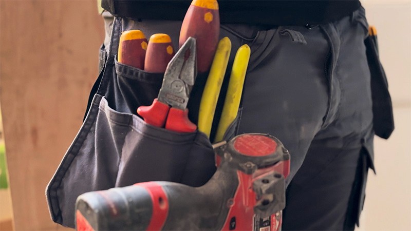 A close-up of a person's hand holding a power tool, with a tool belt and various tools visible.