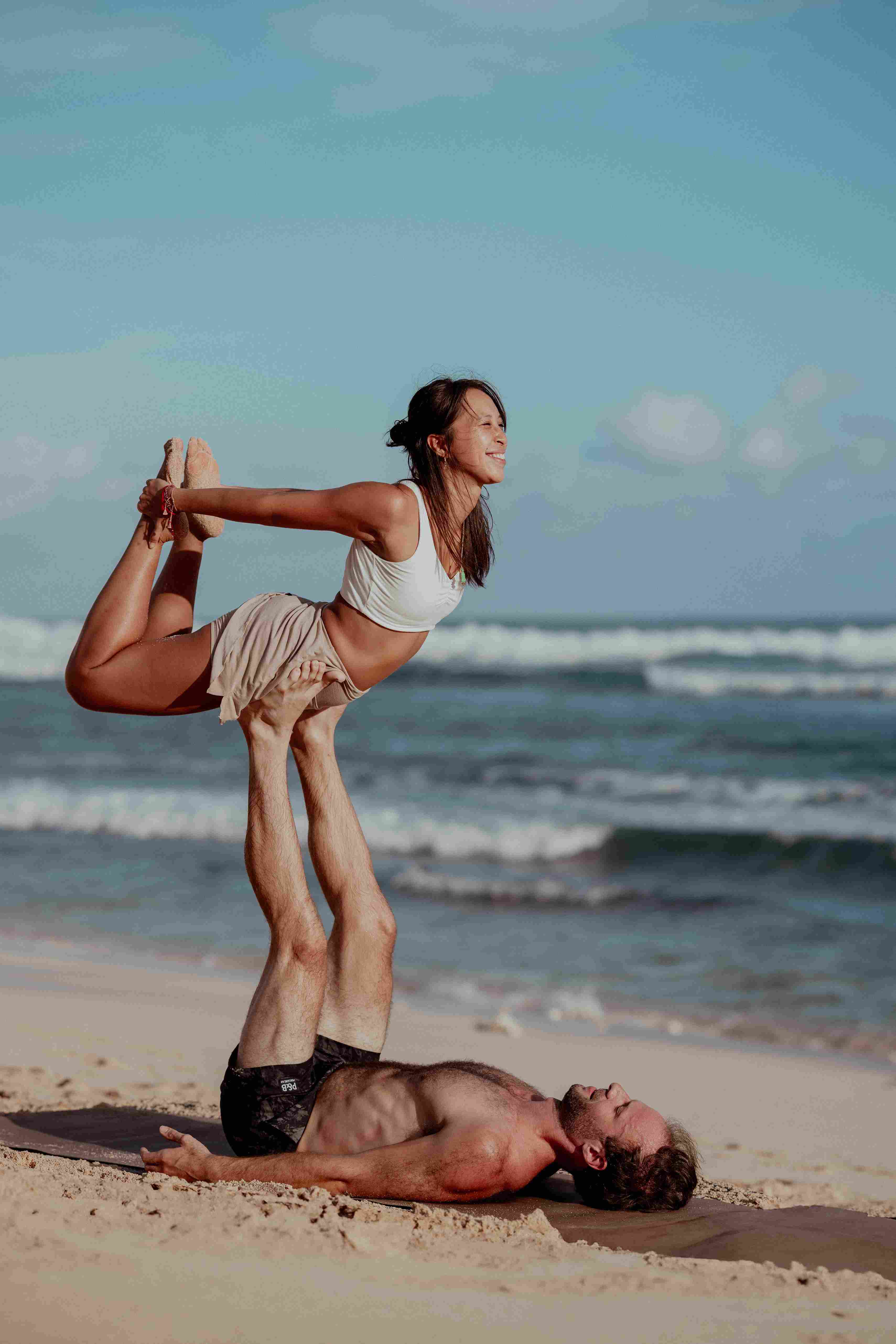 Two students practicing advanced Acro yoga partner bow pose on the beach at sunset.