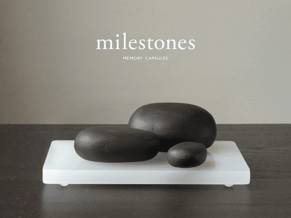Three dark-colored stones rest on a white rectangular base placed on a dark wooden surface. The stones vary in size, with one larger stone and two smaller ones. The background is a light grayish-tan color, and the text "milestones" and "MEMORY CAPSULES" appears in white at the top.