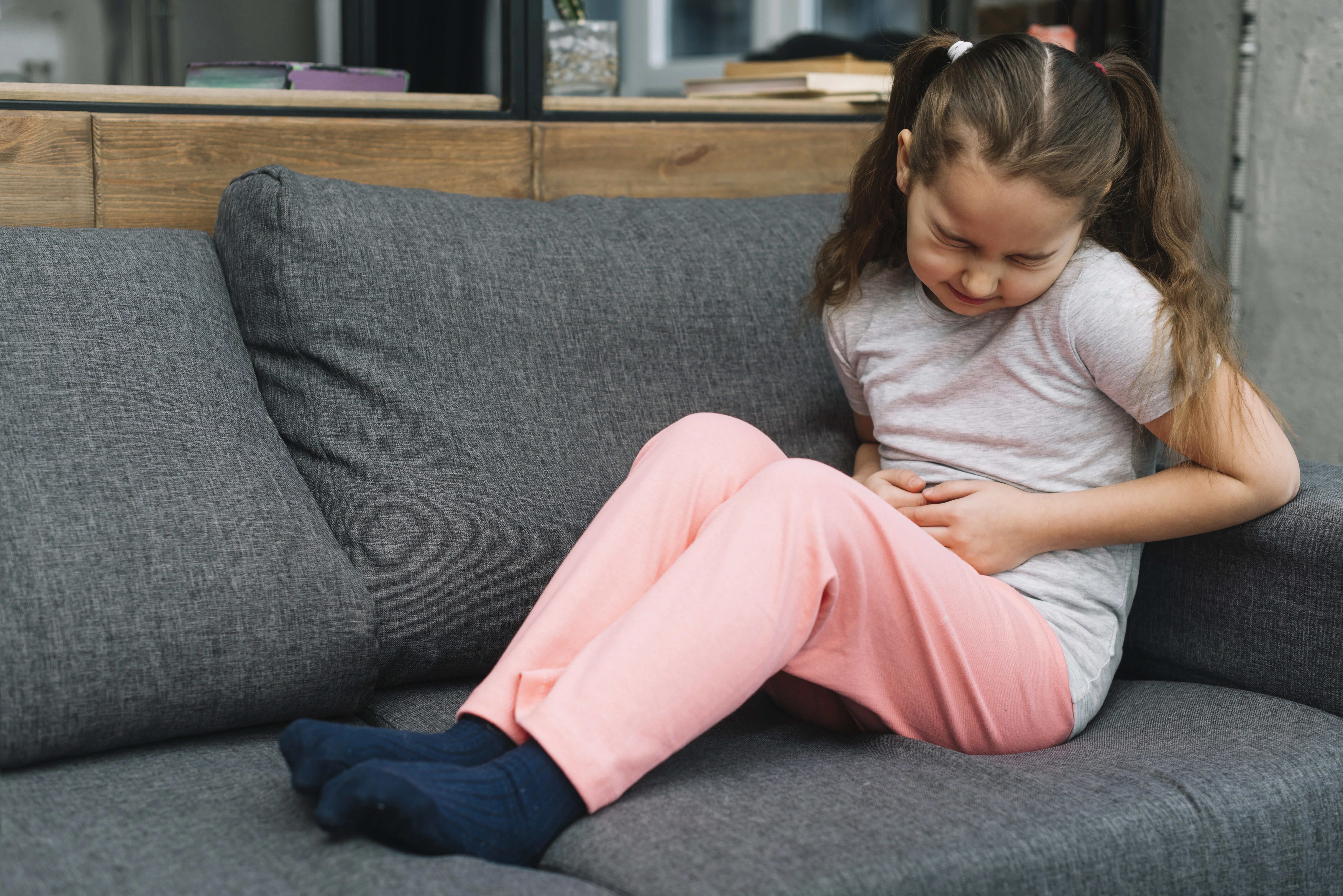 Uma menina com cabelo castanho amarrado em marias-chiquinhas, sentada em um sofá cinza, apertando a barriga com as mãos e fazendo uma expressão de dor ou desconforto abdominal.