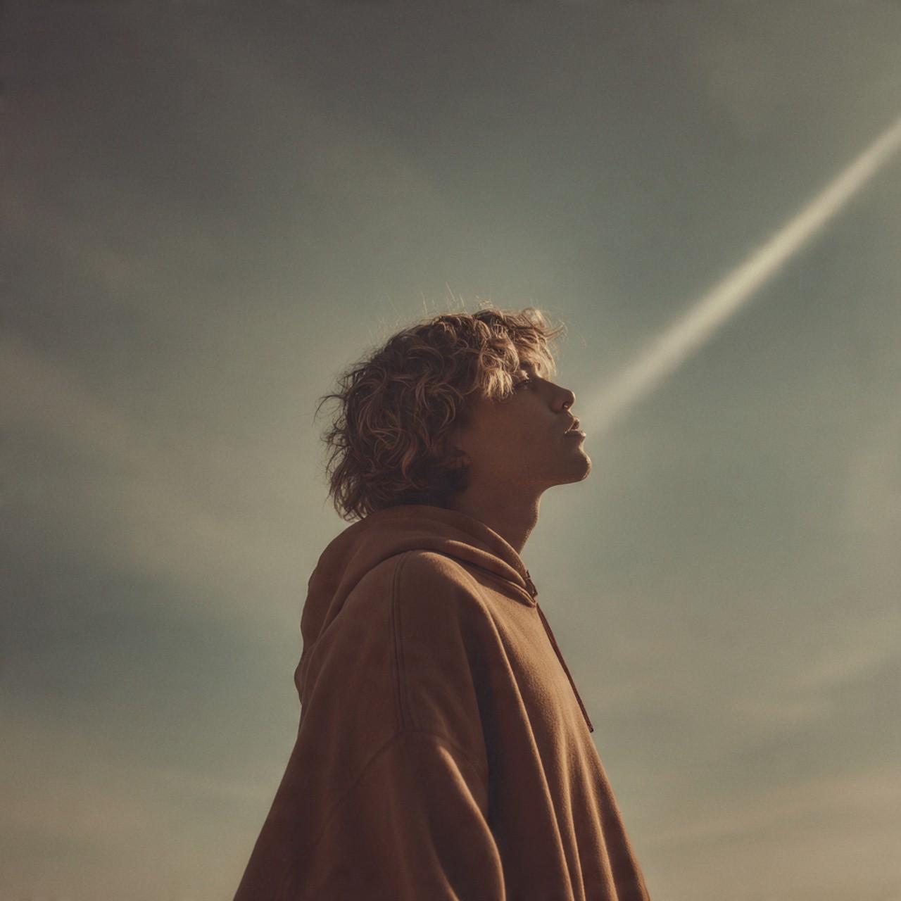 Close-up profile of a young person with curly hair and freckles in a hoodie looking up toward a sunbeam.