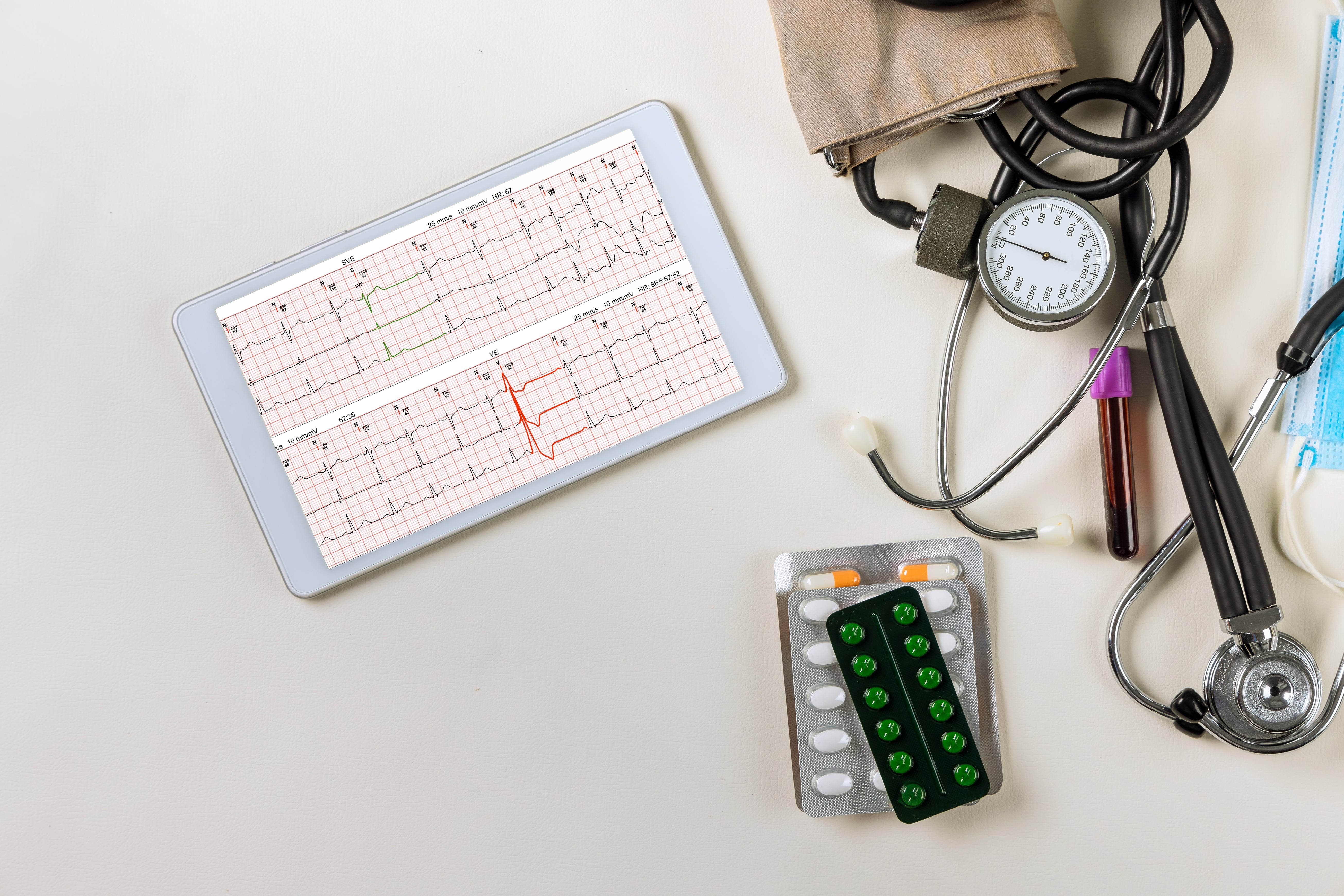Tablet displaying ECG heart monitor data alongside stethoscope, blood pressure monitor, blood sample, pills, and medical face mask on a white surface.